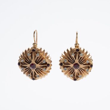 Orecchini pendenti a forma di puff con croce stilizzata, cristallo rosso centrale e cristalli bianchi, su sfondo bianco.