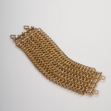 Primo piano dettagliato di un bracciale manchette a catena, in lega francese 'demirouge' dorata 14 KT, su sfondo bianco. Il bracciale presenta una lavorazione a maglia rolò su 5 file e una chiusura a moschettone.