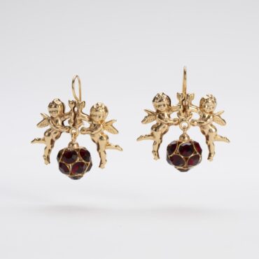 Primo piano dettagliato di un paio di orecchini Cupido in lega demirouge dorata 14 KT con cristalli rossi europei, su sfondo bianco. Gli orecchini mostrano un design elaborato con la figura di Cupido e una boule centrale luminosa.