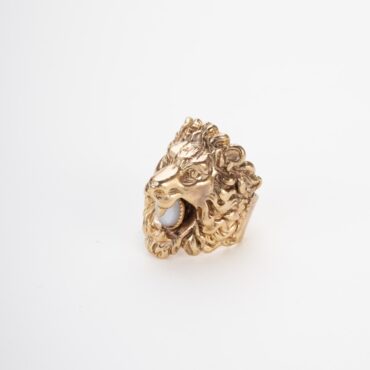 Anello scultoreo dorato con testa di leone in rilievo e pietra dura chiara incastonata nella bocca aperto sul retro su sfondo chiaro.