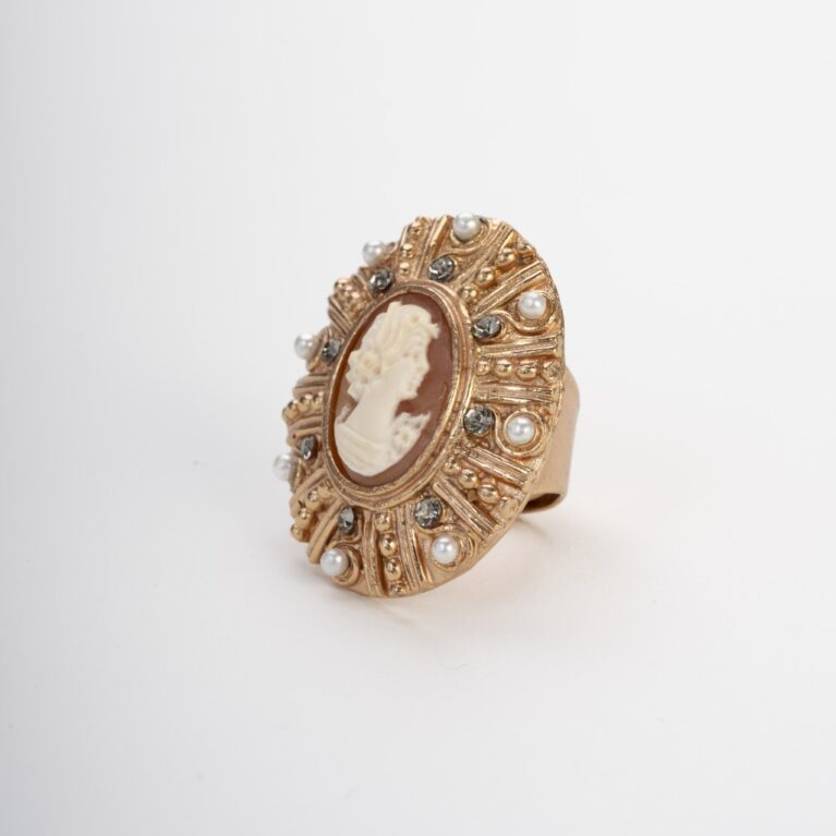Anello ovale artigianale stile vintage con cammeo centrale raffigurante un profilo di donna classica. La montatura è in metallo dorato riccamente lavorata e decorata con piccole perle bianche e pietre grigie incastonate, su sfondo bianco neutro.