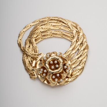 Bracciale multifilo dorato con grande rosone centrale a motivo giglio borbonico e perle su sfondo neutro.