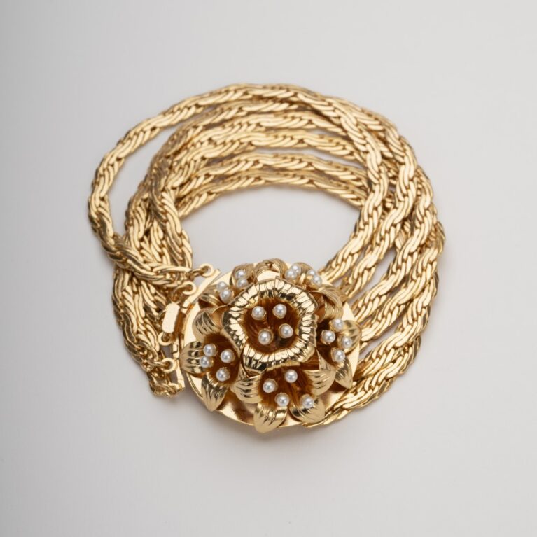 Bracciale multifilo dorato con grande rosone centrale a motivo giglio borbonico e perle su sfondo neutro.