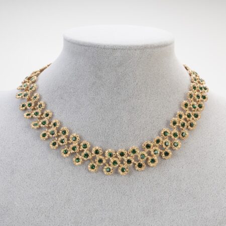 Collier posizionato su busto grigio, mette a fuoco la trama a maglia ricamata, il colore intenso dello smeraldo e la precisione artigianale.