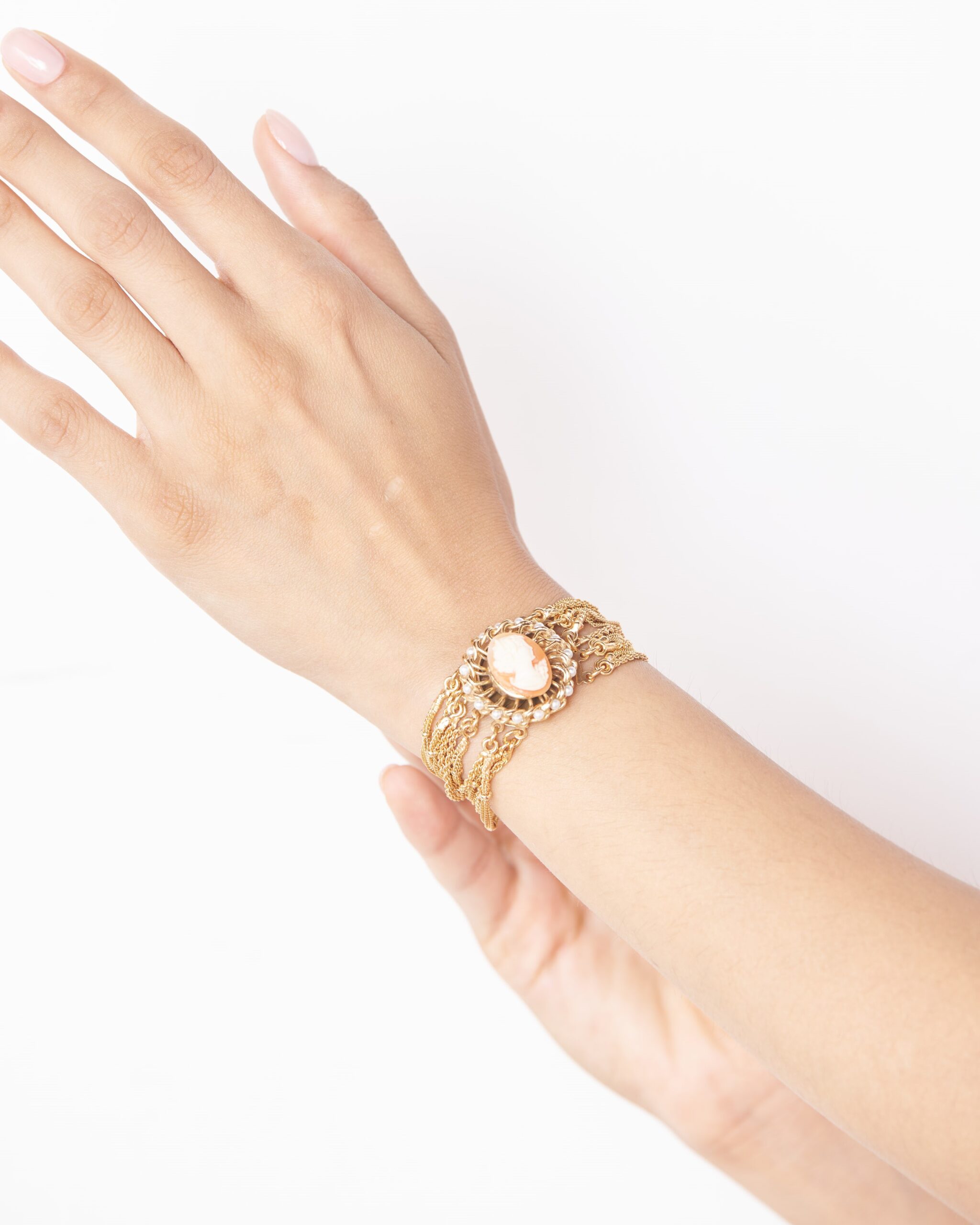 Dettaglio di bracciale scultoreo con cammeo e catene dorate multifilo indossato al polso su sfondo chiaro.